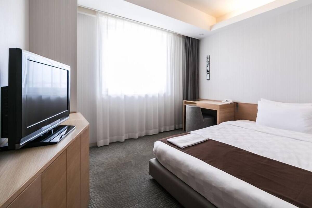 A hotel room with a bed, a TV on a wooden stand, a desk with a chair, and a window with curtains.