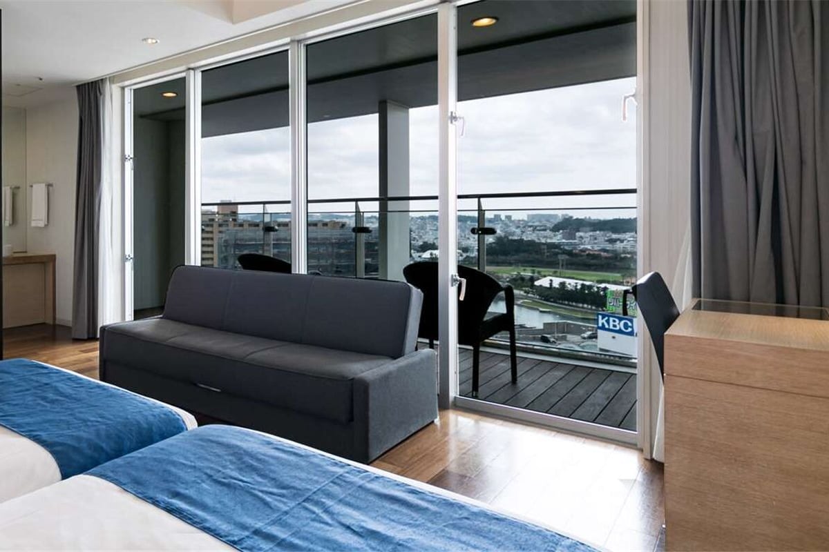 A modern hotel room with a large TV, a sofa, a bed, and a balcony with a city view.