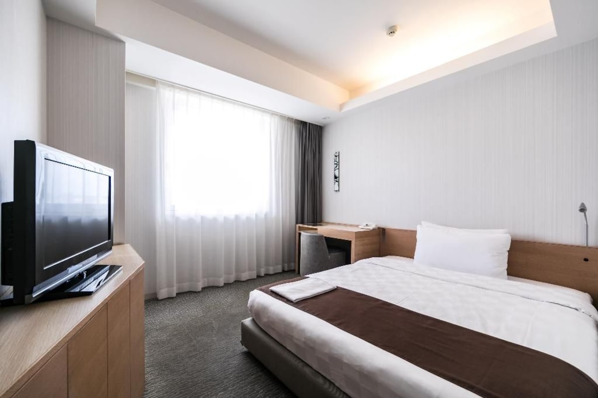 A hotel room with a bed, a desk, a television, and a window with curtains.