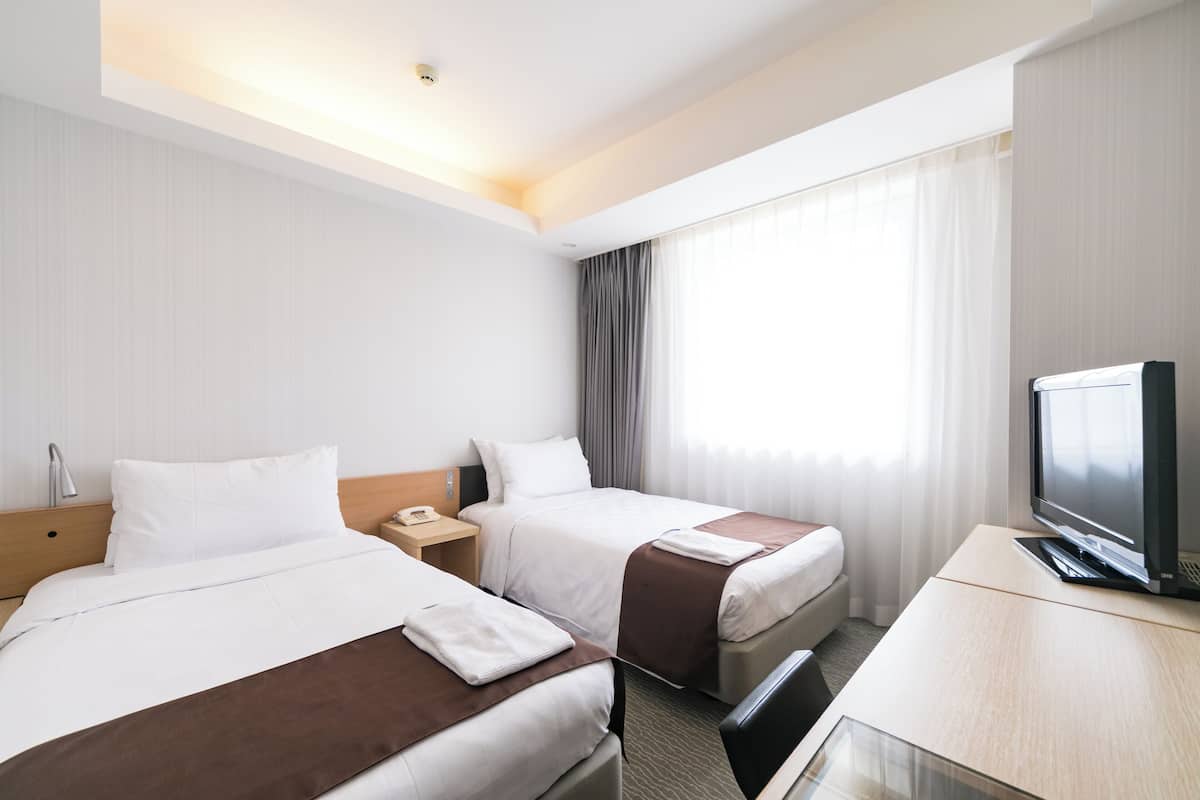 A hotel room with two beds, a TV, a desk, and a window with curtains.