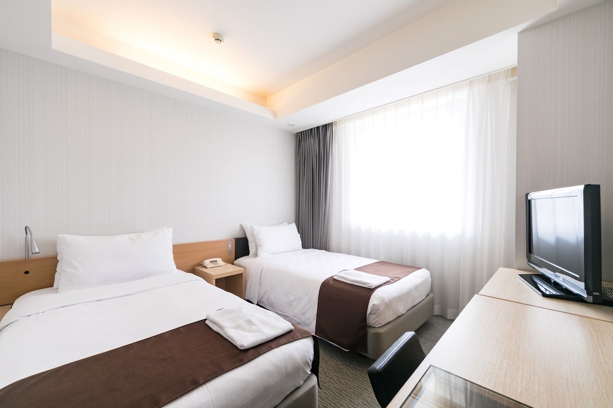A hotel room with two beds, a desk with a television, a telephone, and a window with curtains.