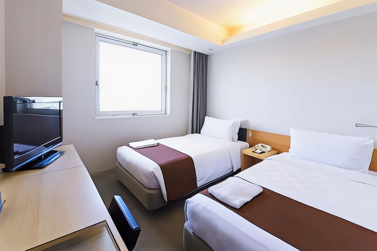 A hotel room with two beds, a desk with a television, a window, and a nightstand with a phone.