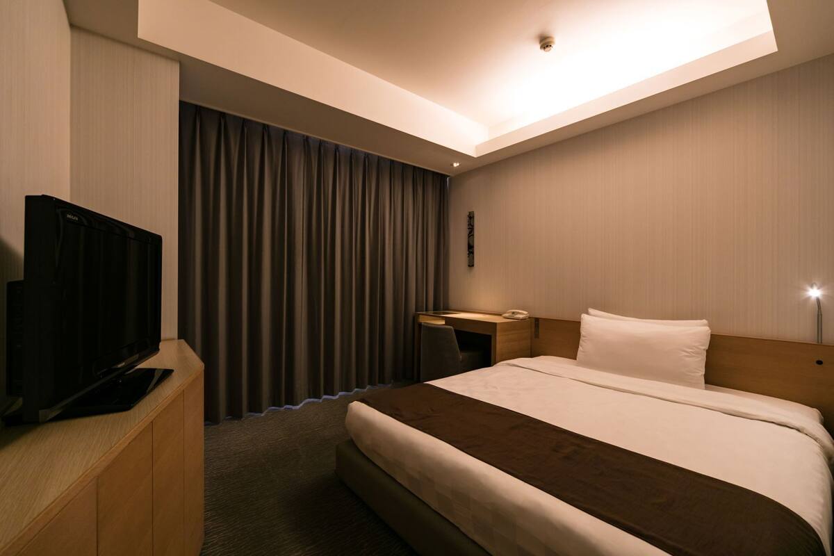 A hotel room with a bed, a desk with a chair, a television, and a curtain.