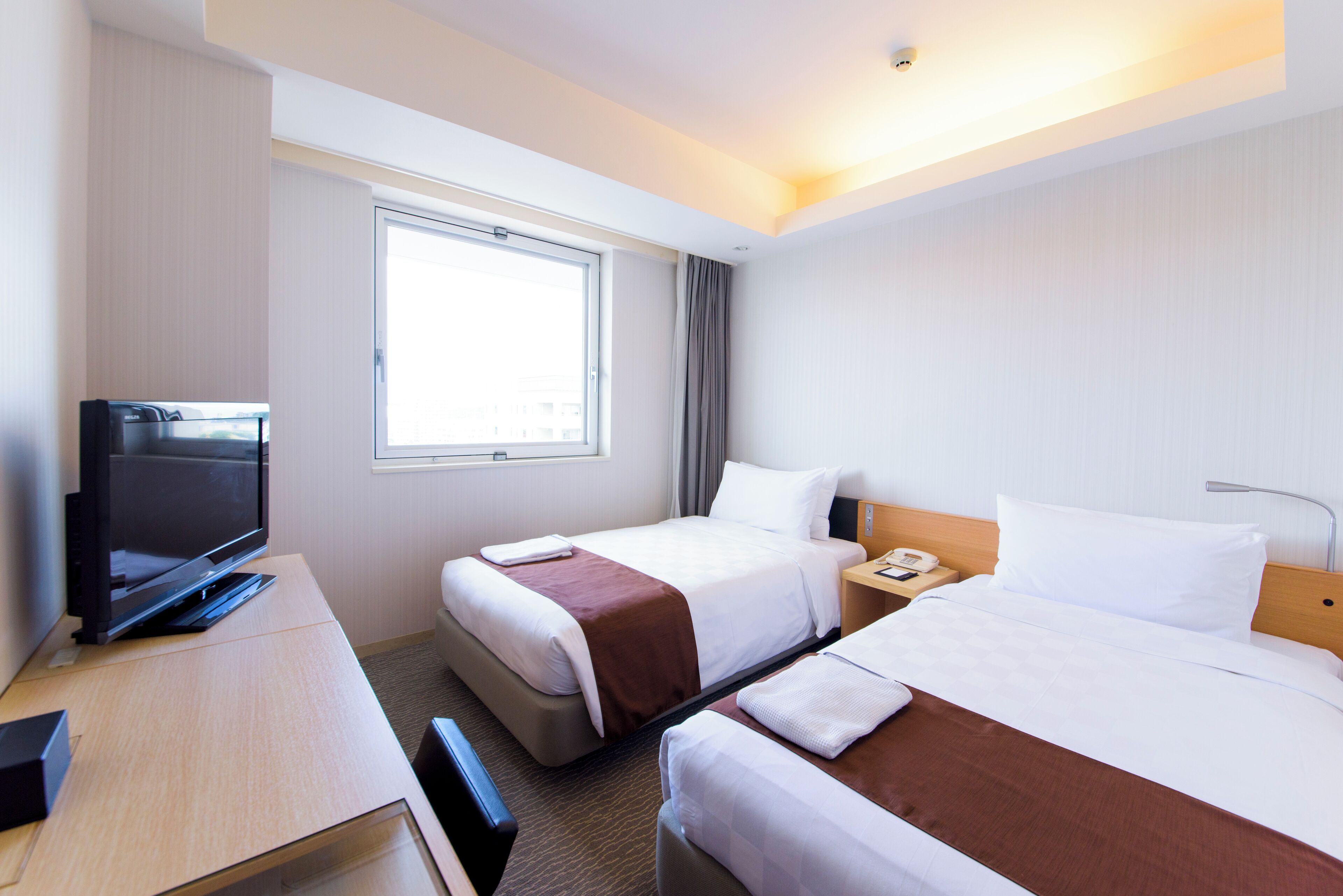 A hotel room with two beds, a desk with a television, a window with curtains, and a bedside table with a phone.