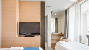 A modern hotel room with a flat-screen TV mounted on the wall, a large bed with a blue accent, a separate bed, a bathroom with a sink and mirror, and a view of the city through large windows. - Naha Tokyu REI Hotel (Naha)