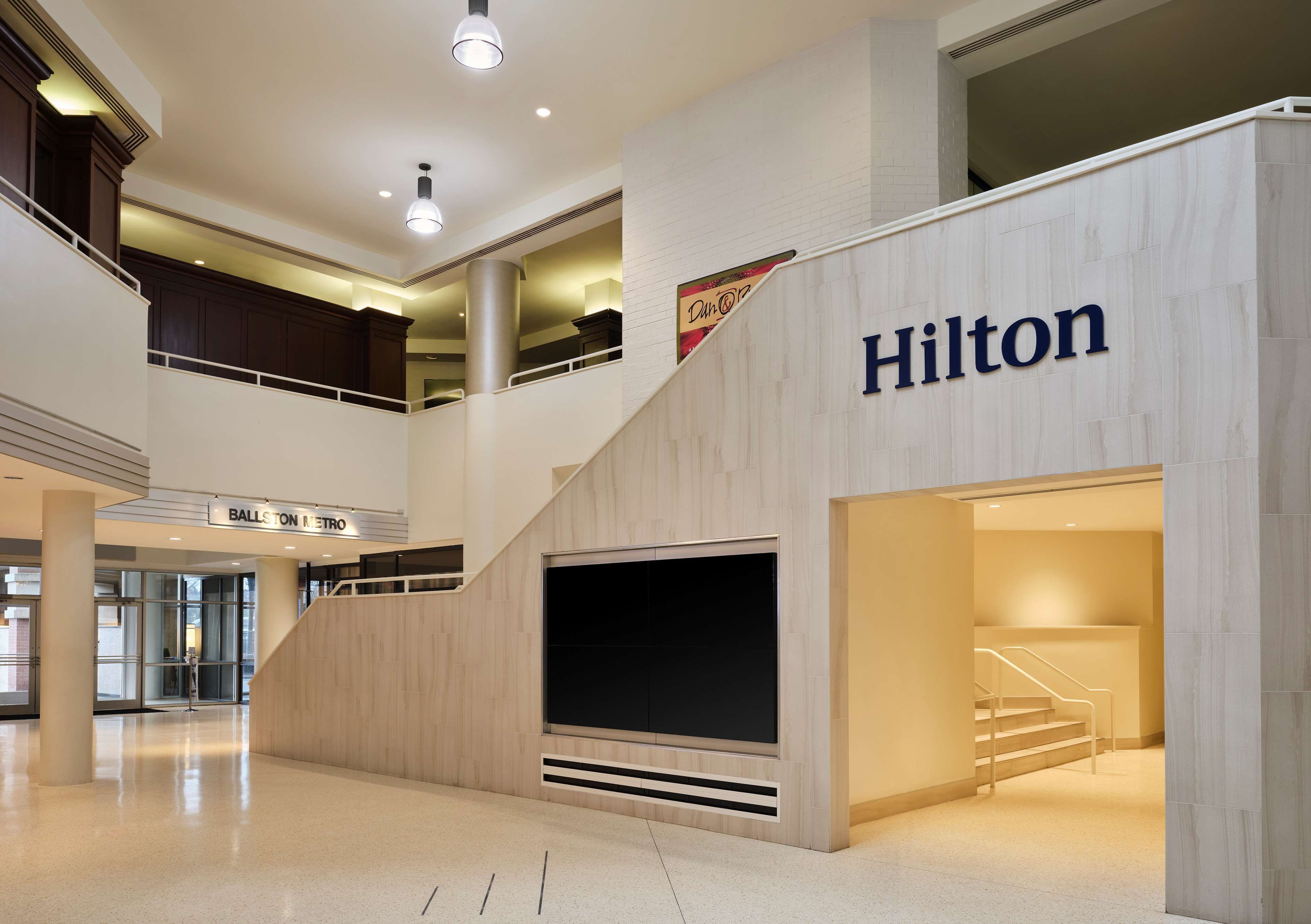 A modern hotel interior with a prominent 'Hilton' sign, a staircase, and a 'Ballston Metro' sign indicating proximity to public transport.