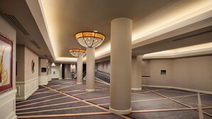 Un couloir d'hôtel avec un sol recouvert de moquette, des colonnes et des lampes suspendues.