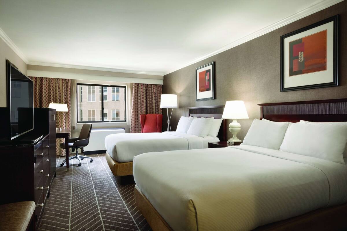 A hotel room with two beds, a desk with a TV, a chair, and a window with curtains.