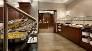 Un espace buffet comprenant plusieurs présentoirs de nourriture, un comptoir en bois et donnant sur un couloir menant à une pièce dotée d’un grand miroir.