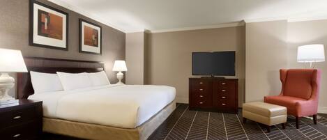 A hotel room with a large bed, a television, a desk with a lamp, a chair, and a small ottoman.