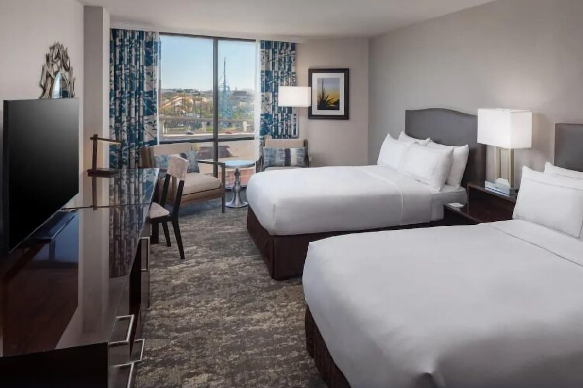 A hotel room with two beds, a desk, a TV, and a view of the city.
