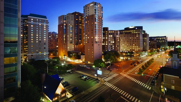 Une vue sur la ville au crépuscule, avec des gratte-ciel illuminés, une intersection bien éclairée et une zone de stationnement.