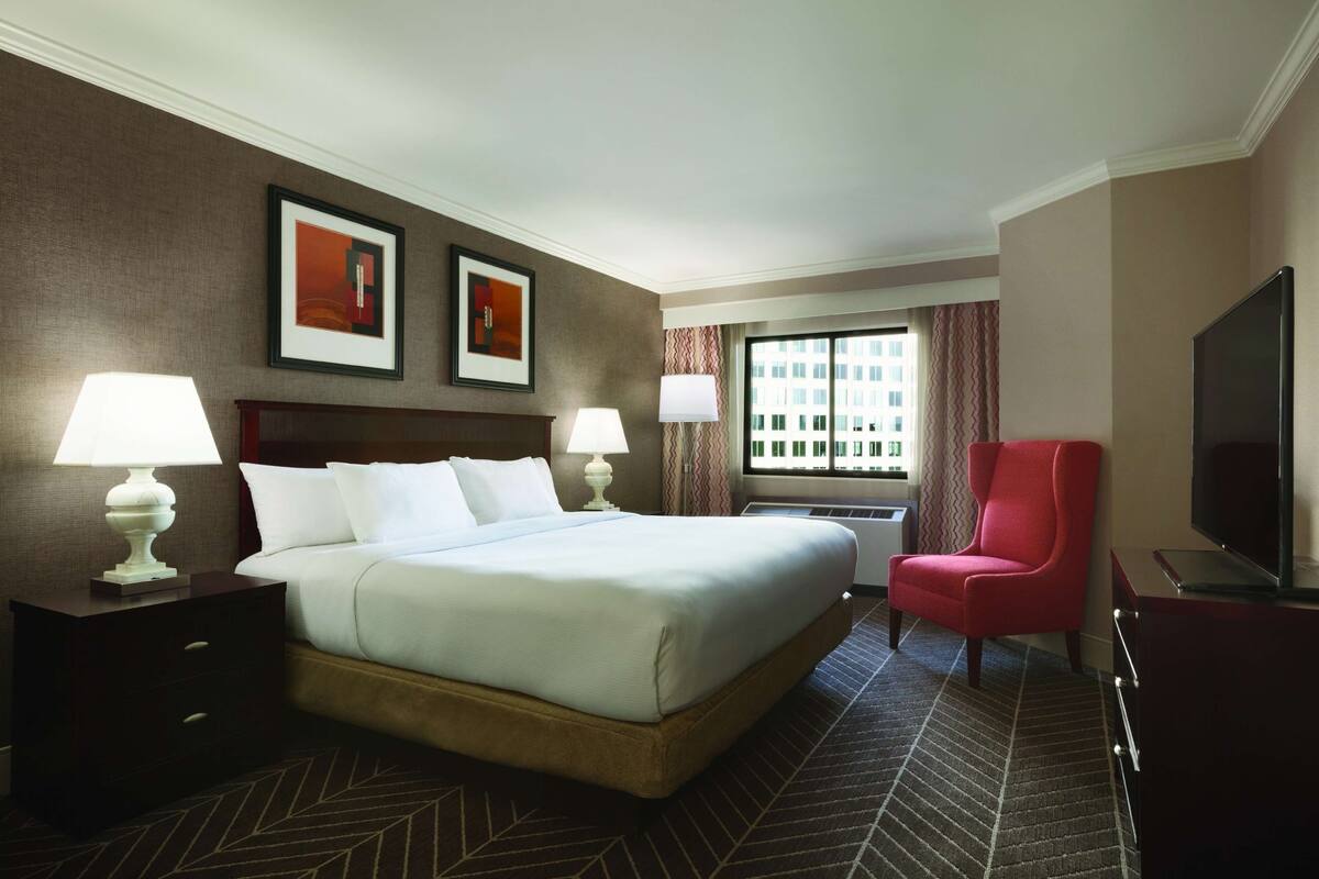 A hotel room with a large bed, two bedside tables with lamps, a red armchair, a TV, and a window with curtains.