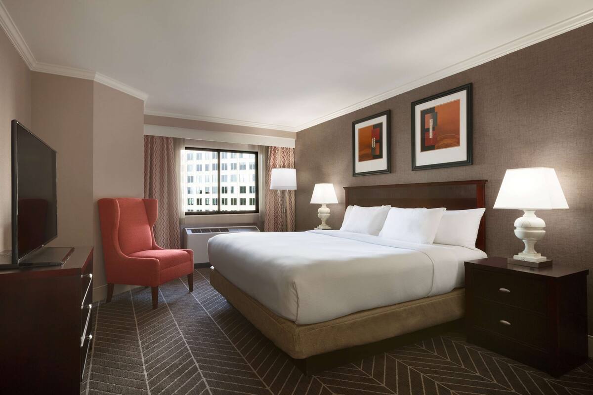 A hotel room with a large bed, two bedside lamps, a TV, a red chair, and a window with curtains.