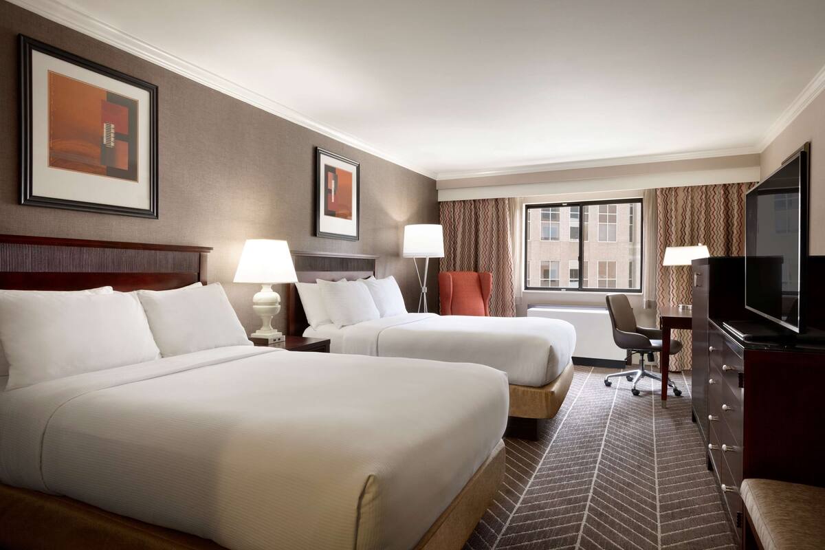A hotel room with two beds, a desk, a chair, a TV, and a window with curtains.