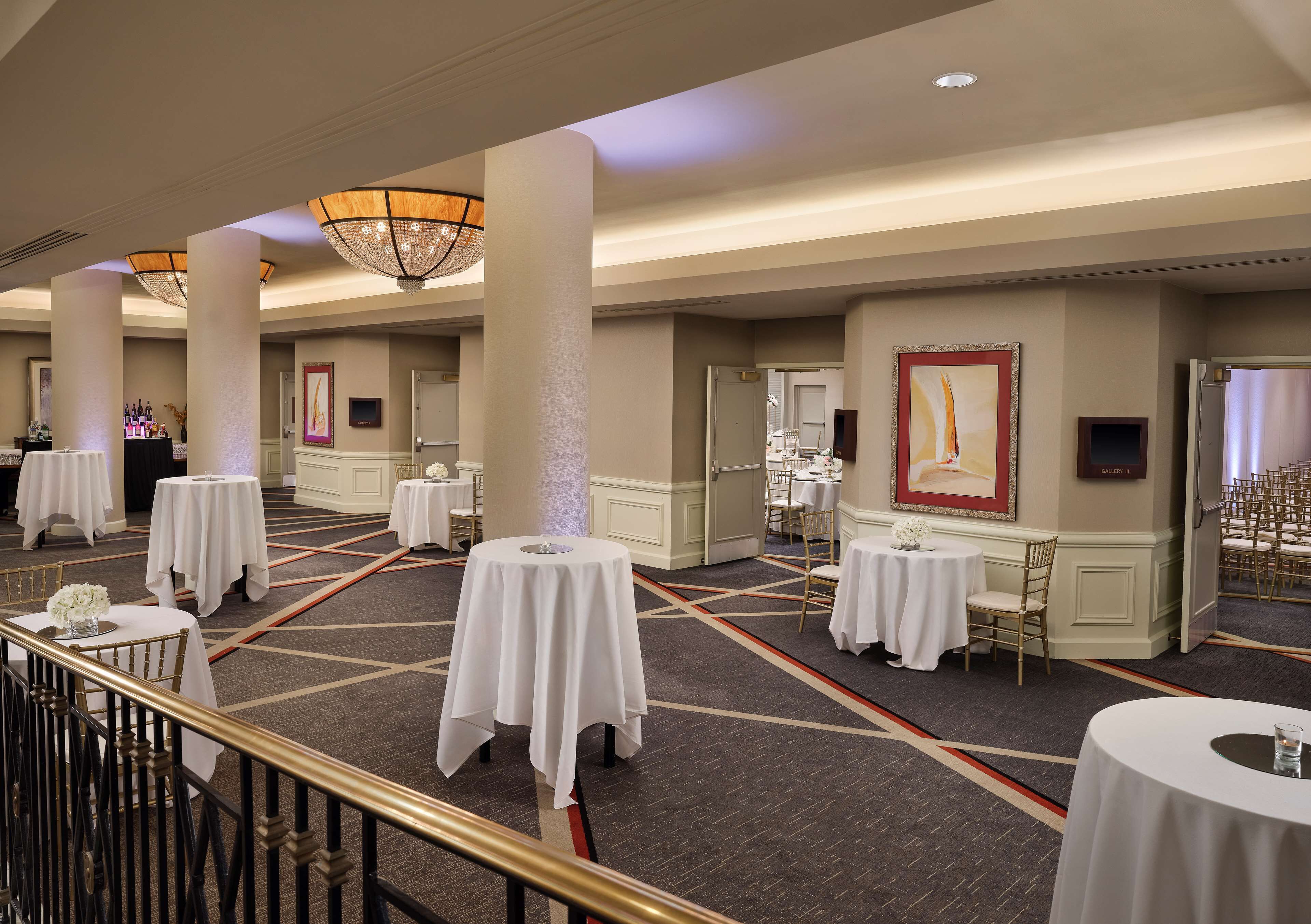 A hotel ballroom with multiple round tables covered with white tablecloths, large columns, and a chandelier.