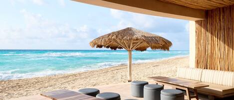 A beachside lounge with a thatched umbrella, wooden tables, and cushioned seating.