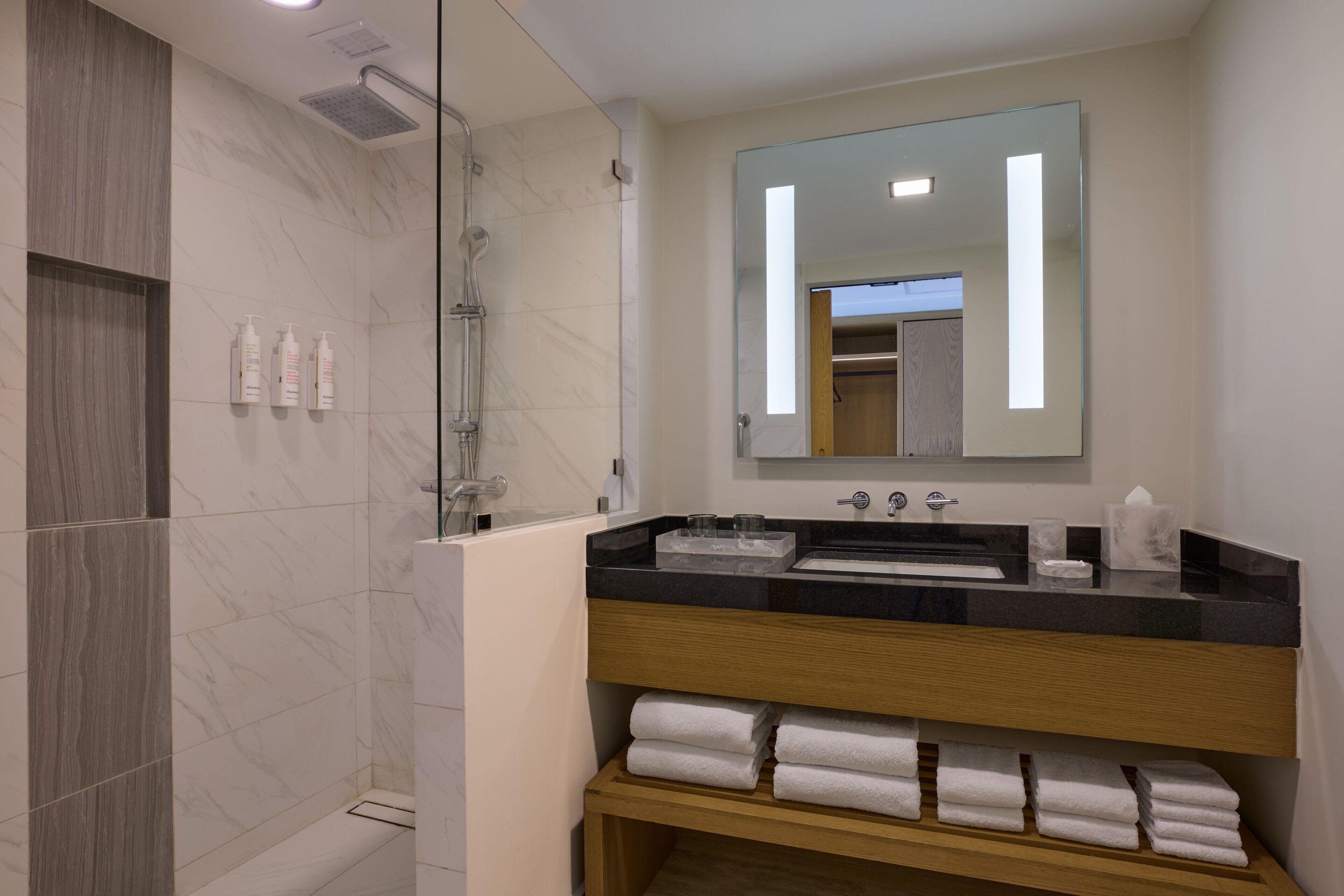 A modern bathroom with a glass-enclosed shower, a black countertop sink, and a mirror above it. There are white towels neatly arranged on a shelf below the sink.