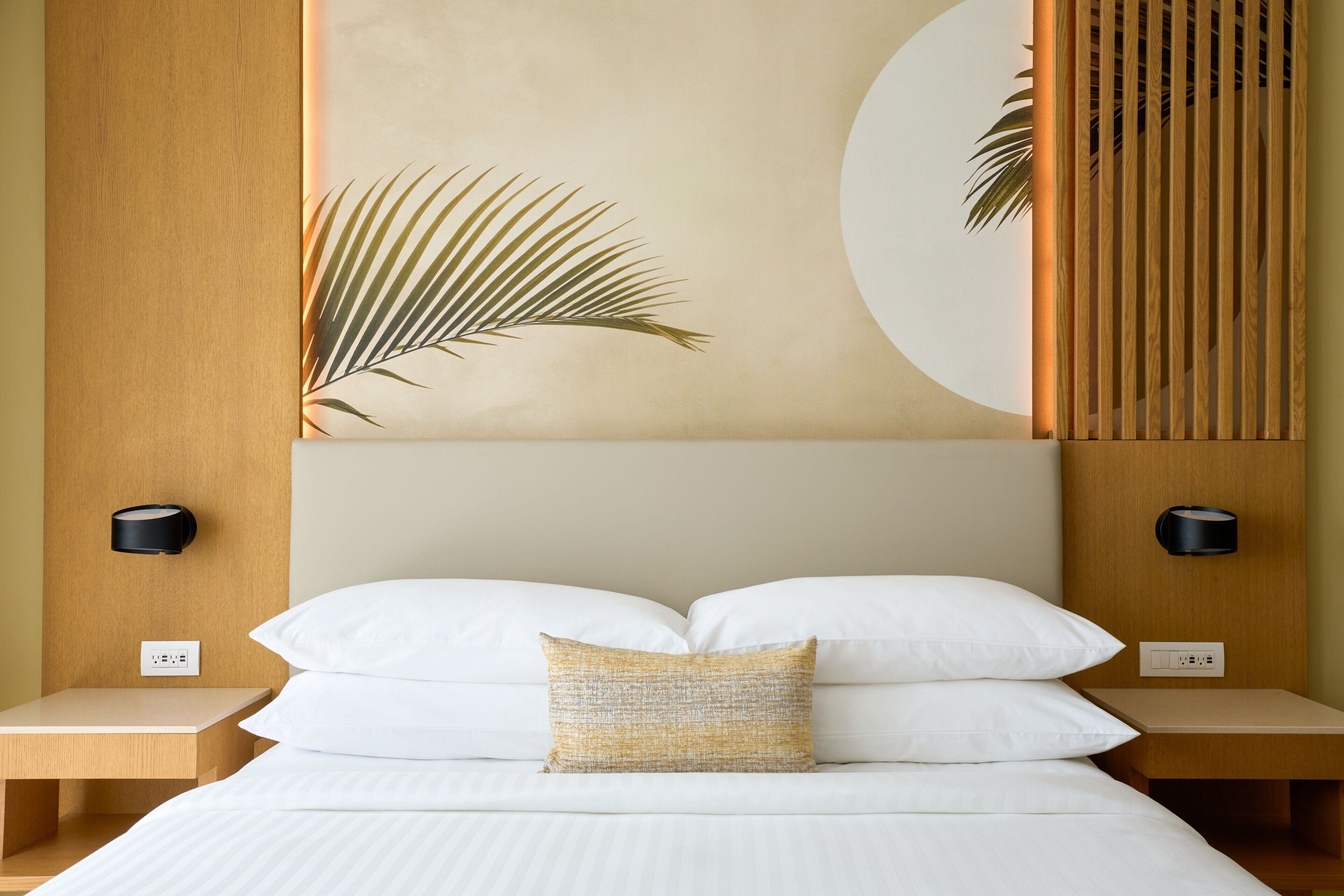 Ein ordentlich eingerichtetes Hotelzimmer mit Bett, weißen Leinentextilien, einem dekorativen Kissen und einem an der Wand befestigten Nachttisch mit Steckdose.