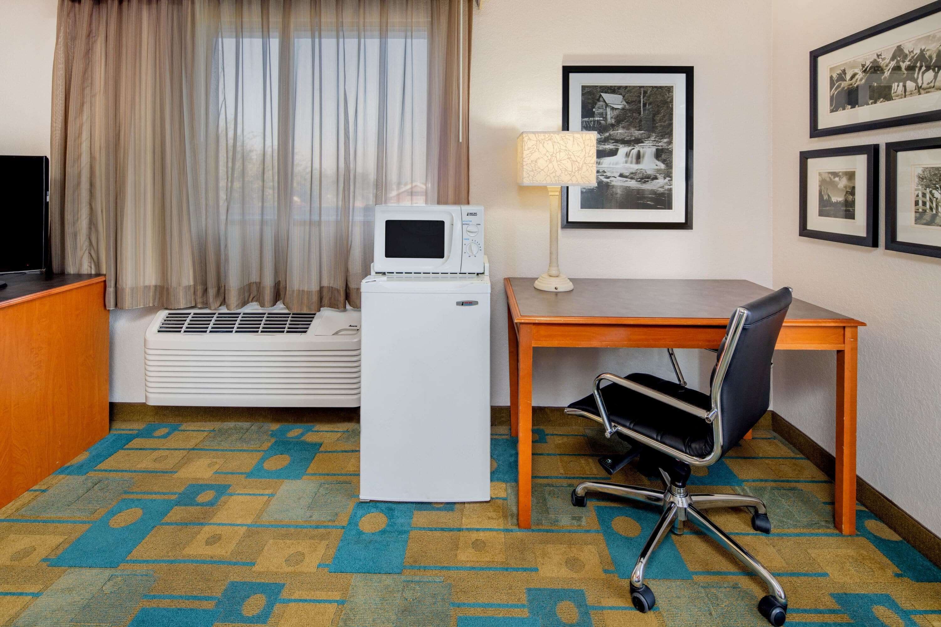 A hotel room with a bed, a desk with a chair, a microwave, a lamp, and framed pictures on the wall.