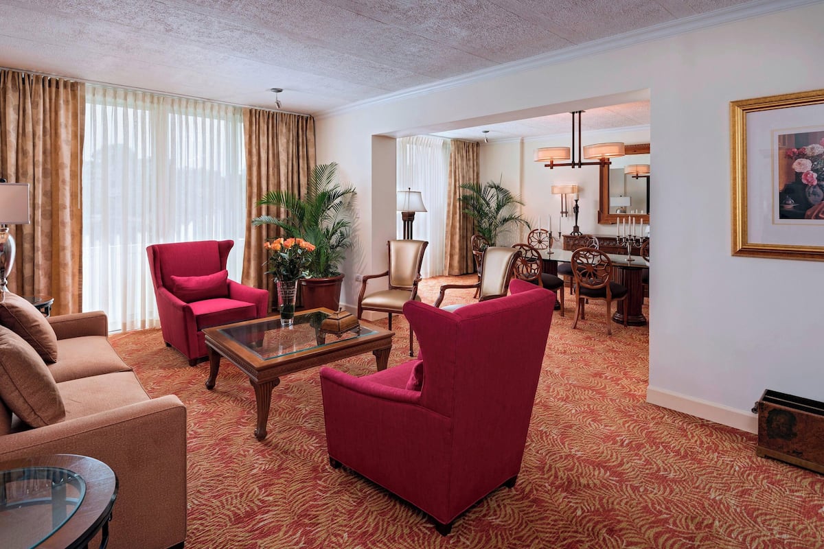 A hotel room with a living area featuring a red armchair, beige sofa, and a glass coffee table. There is a dining area with a wooden table and chairs, and a large mirror on the wall.