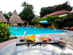 Una piscina con sillas de descanso, una mesa con comida y estructuras de techo de paja.
