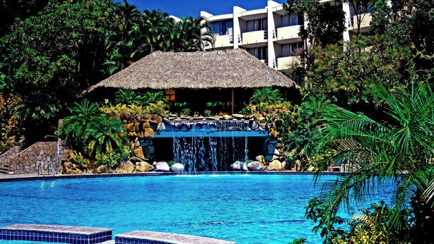 Una piscina con cascada, rodeada de follaje tropical y una estructura con techo de paja.
