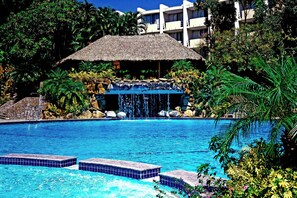 Una piscina con cascada, rodeada de follaje tropical y una estructura con techo de paja.