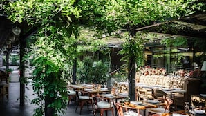 An outdoor dining area with wooden tables and chairs, surrounded by lush greenery and a glass wall. - J House Greenwich (Riverside)
