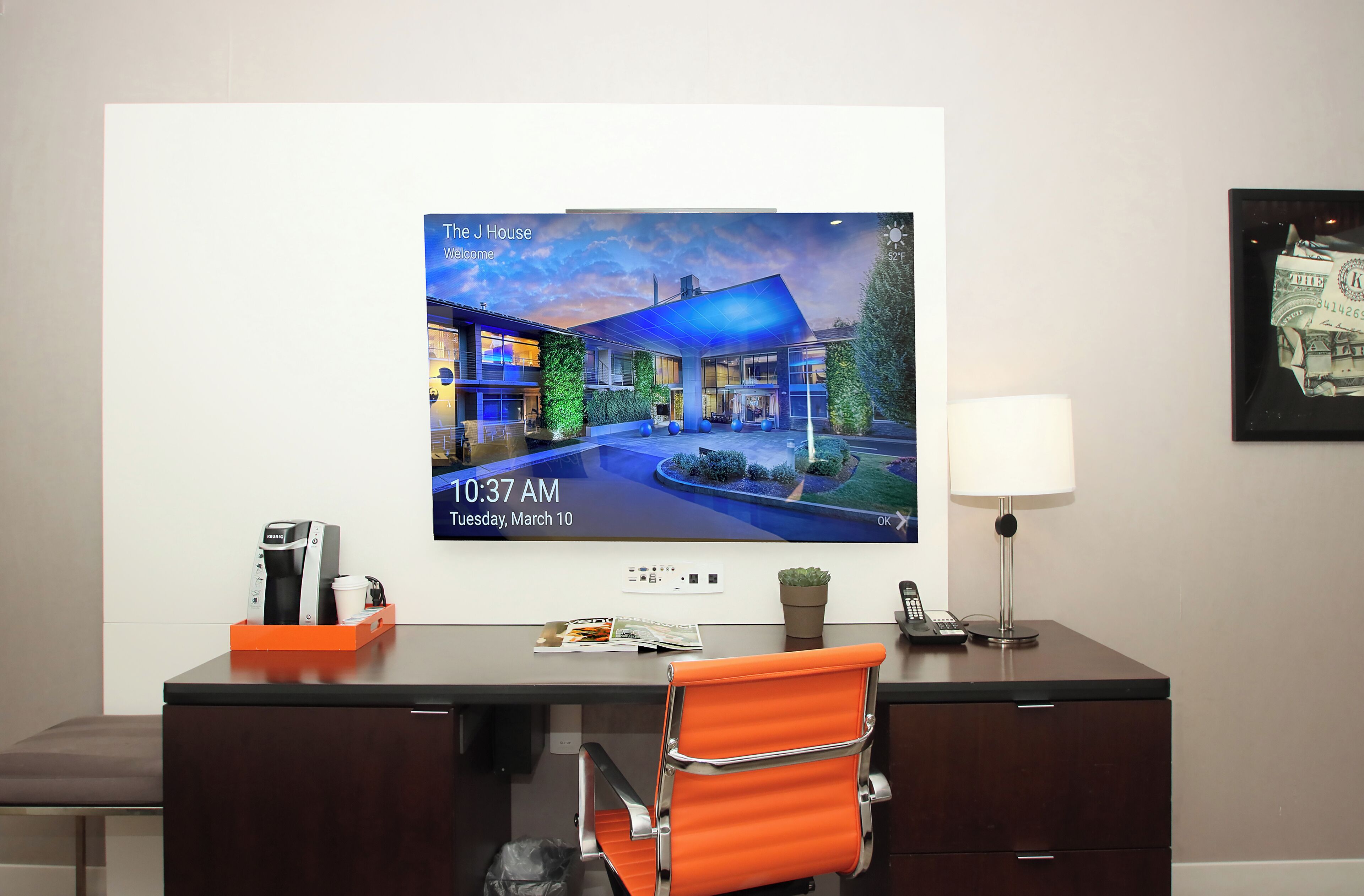 Habitación de hotel con televisión mostrando una imagen del establecimiento, escritorio con cafetera, lámpara y una silla.