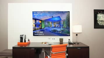 Hotellihuone, jossa on televisio, jolla näkyy kuva hotellista, työpöytä, kahvinkeitin, lamppu ja tuoli.