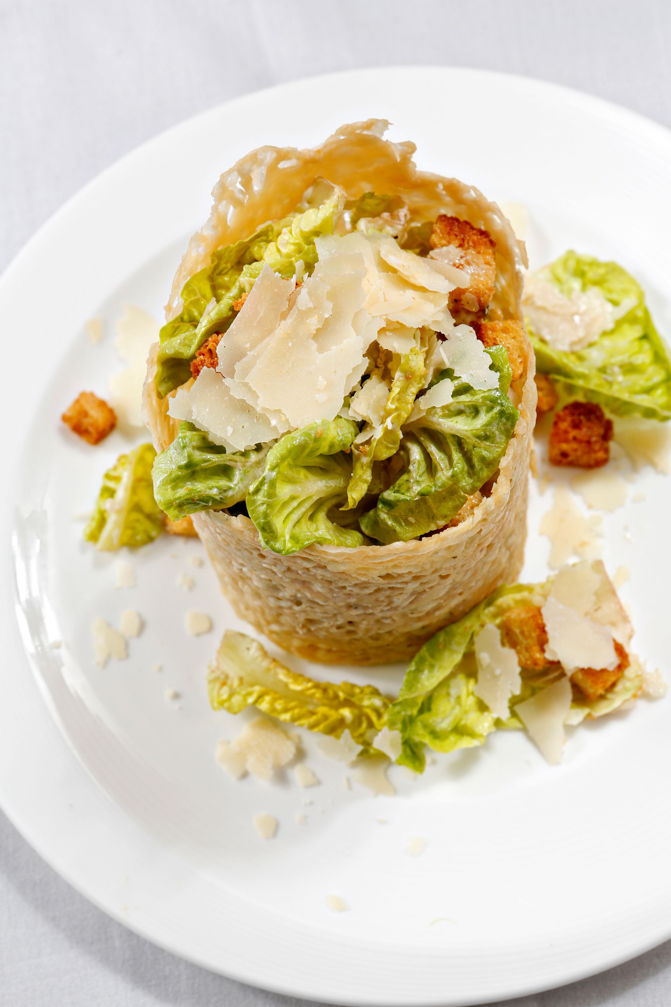 Un plato blanco con un alimento envuelto, posiblemente una ensalada o aperitivo, adornado con queso rallado y crutones.