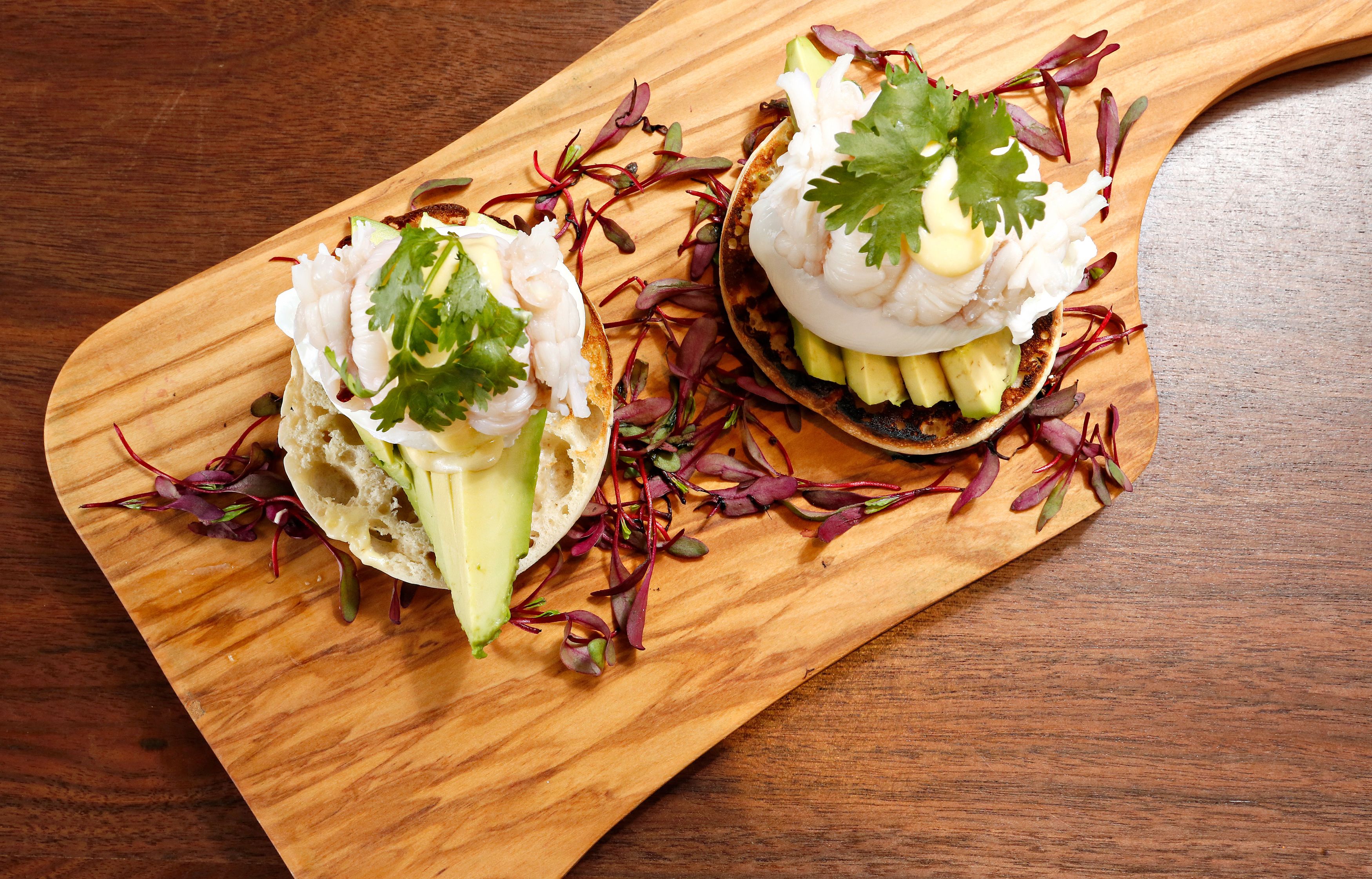 Two tacos on a wooden board with avocado, cilantro, and a creamy topping, surrounded by red and purple leaves.