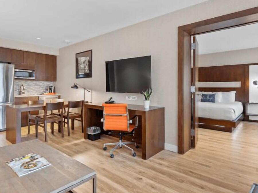 A modern hotel room with a wooden floor, a desk with a chair, a bed, a TV, and a kitchen area.