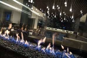 A modern interior with a fire feature on a pebble-lined surface, contemporary furniture, and a high ceiling with hanging lights. - DoubleTree by Hilton Hotel Syracuse (East Syracuse)