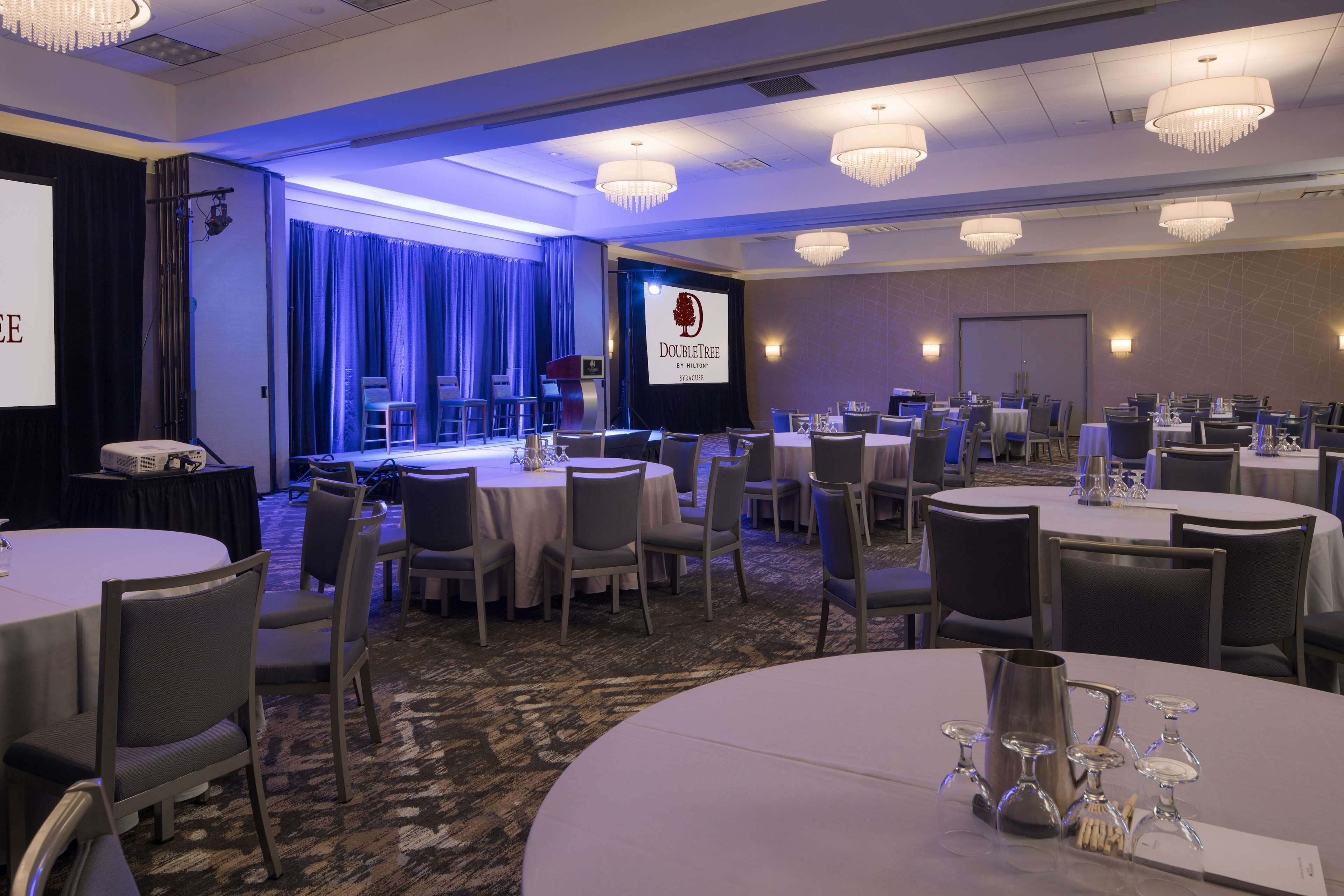 A conference room with multiple round tables, chairs, and a stage with a screen displaying "DoubleTree by Hilton".