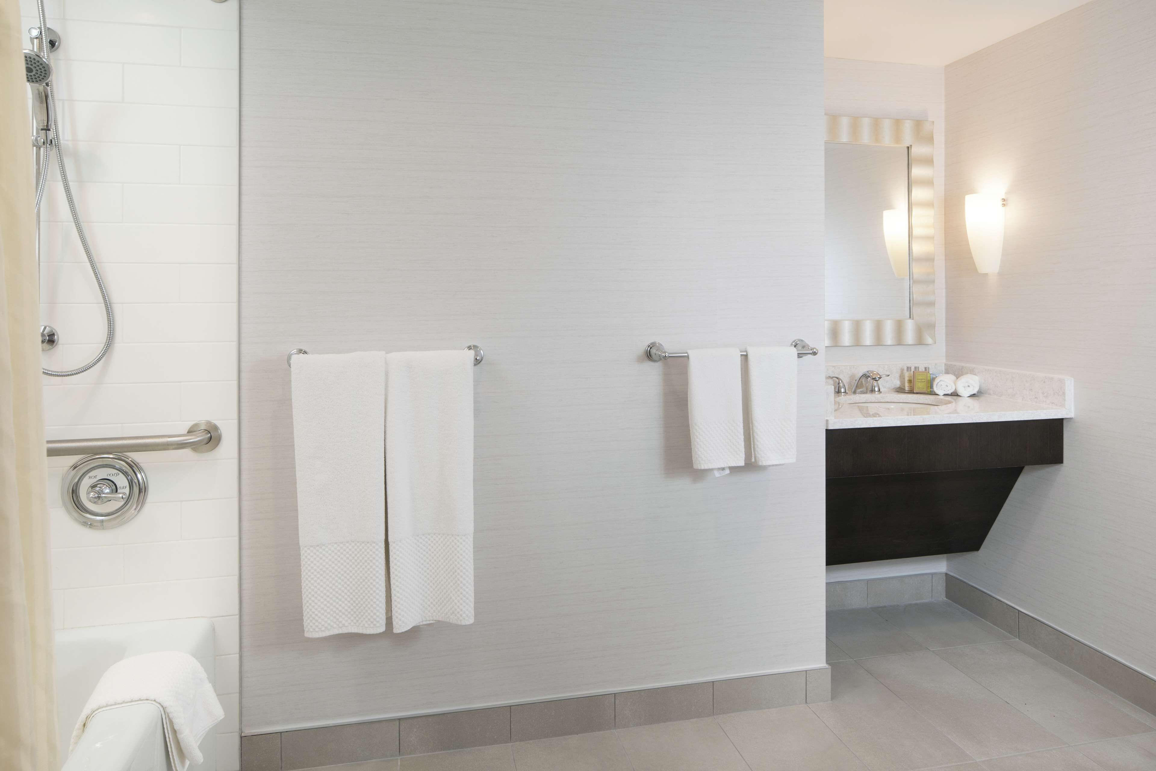 Un baño moderno con ducha, dos toallas colgadas en la pared, lavamanos con espejo e iluminación.