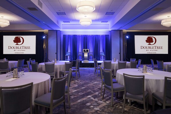 Une salle de conférence avec des tables et des chaises rondes, une scène avec un rideau bleu en arrière-plan et un grand écran affichant le logo de DoubleTree by Hilton.