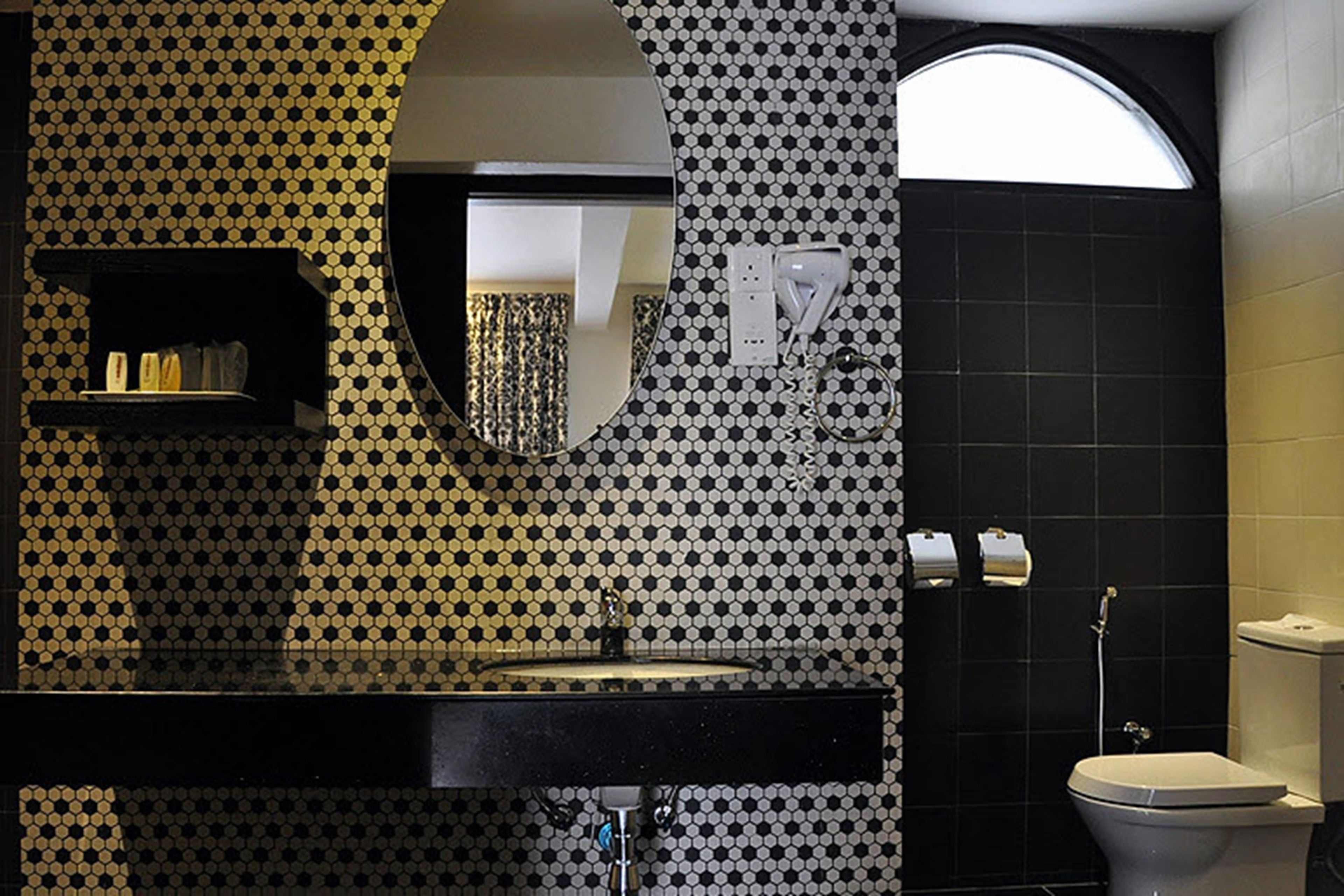 A bathroom with a black and white checkered wall pattern, a round mirror, a black sink, a toilet, and a towel dispenser.