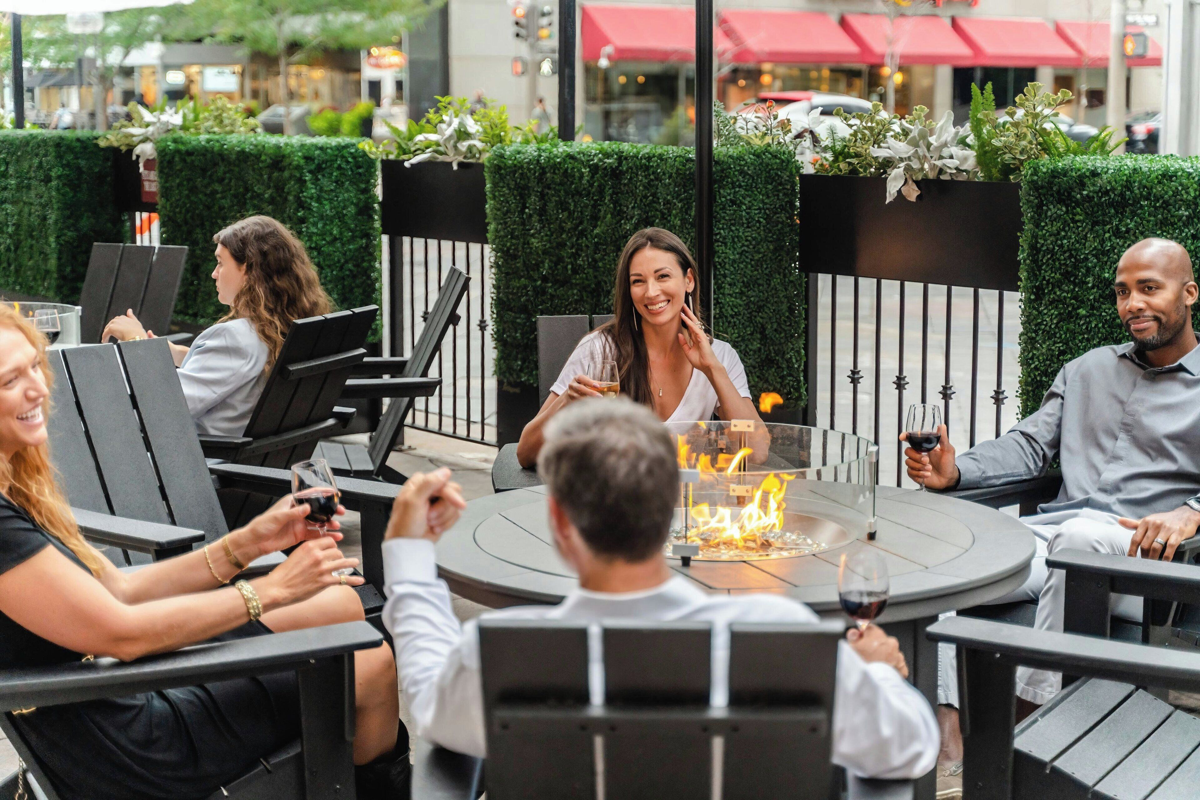 Un groupe de personnes est assis autour d’une table ronde installée à l’extérieur, près d’un foyer, dans un cadre urbain en plein air agrémenté de verdure et d’une haie en arrière-plan.