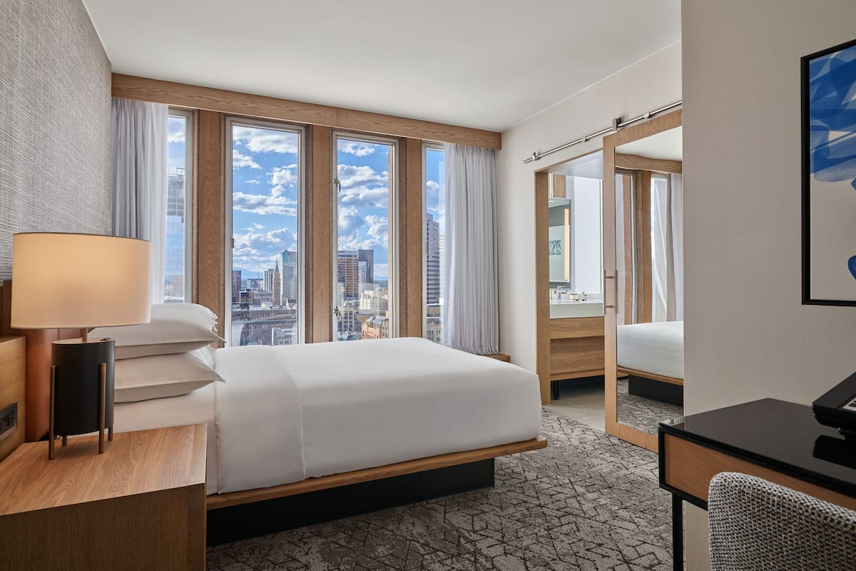 A hotel room with a large bed, a desk, a chair, and a view of the city through large windows.