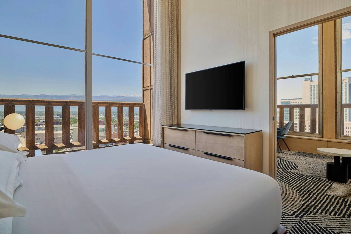 A hotel room with a large bed, a flat-screen TV mounted on the wall, and a balcony with a view of the city and mountains.