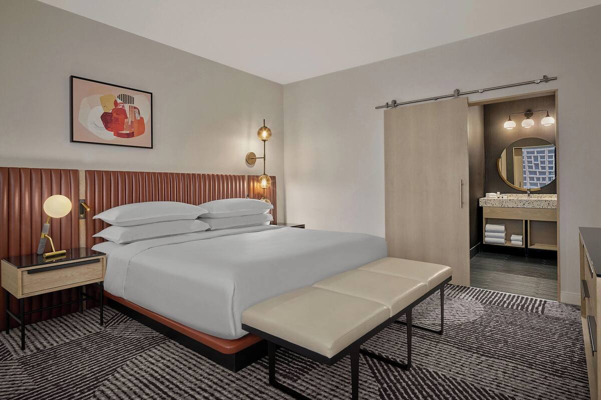 A hotel room with a bed, bedside table, bench, and a bathroom with a mirror and sink.
