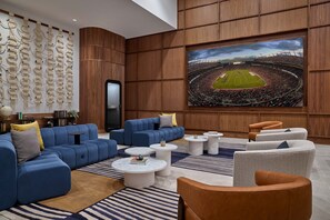 A modern lounge with a large screen displaying a stadium, blue sofas, and round coffee tables. - Sheraton Denver Downtown Hotel (Denver)