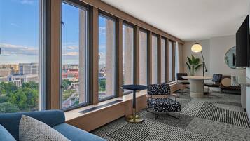 A modern hotel room with large windows, a sofa, a small round table, a desk, and a TV.