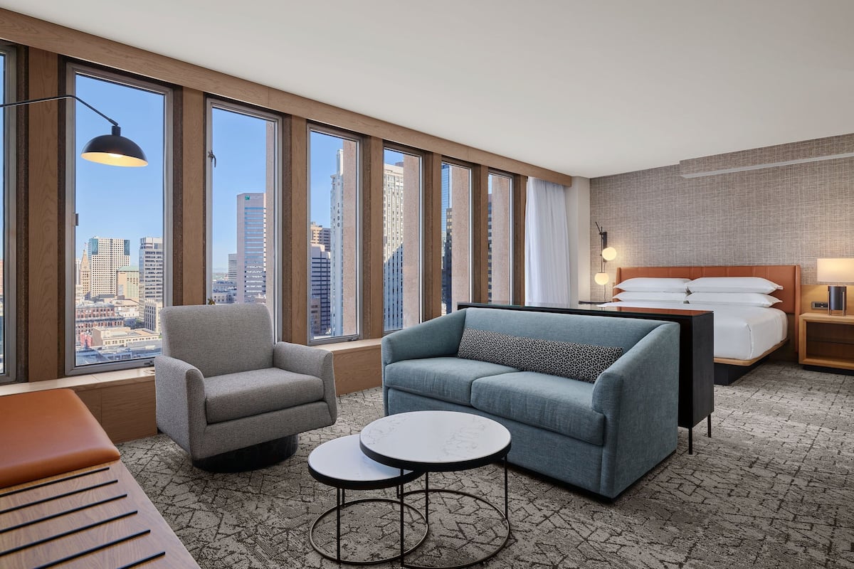 A hotel room with a large window, a bed, a sofa, a chair, and a small table.