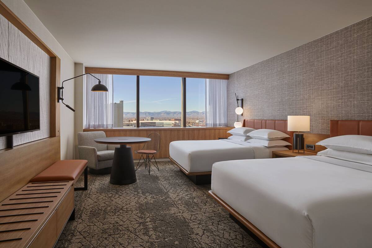 A hotel room with two beds, a desk, a chair, a TV, and a large window with a city view.