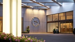 A modern hotel entrance with a large, illuminated logo on the wall, large windows, and potted plants.