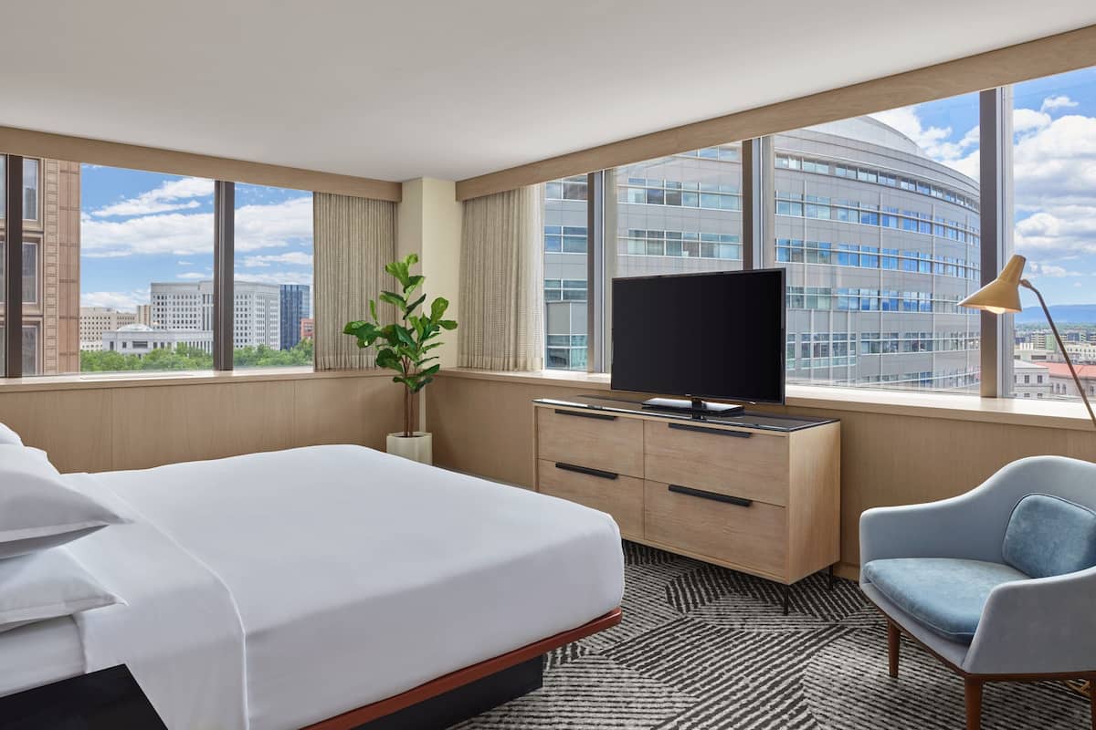 A hotel room with a bed, a desk with a TV, a chair, and large windows offering a city view.