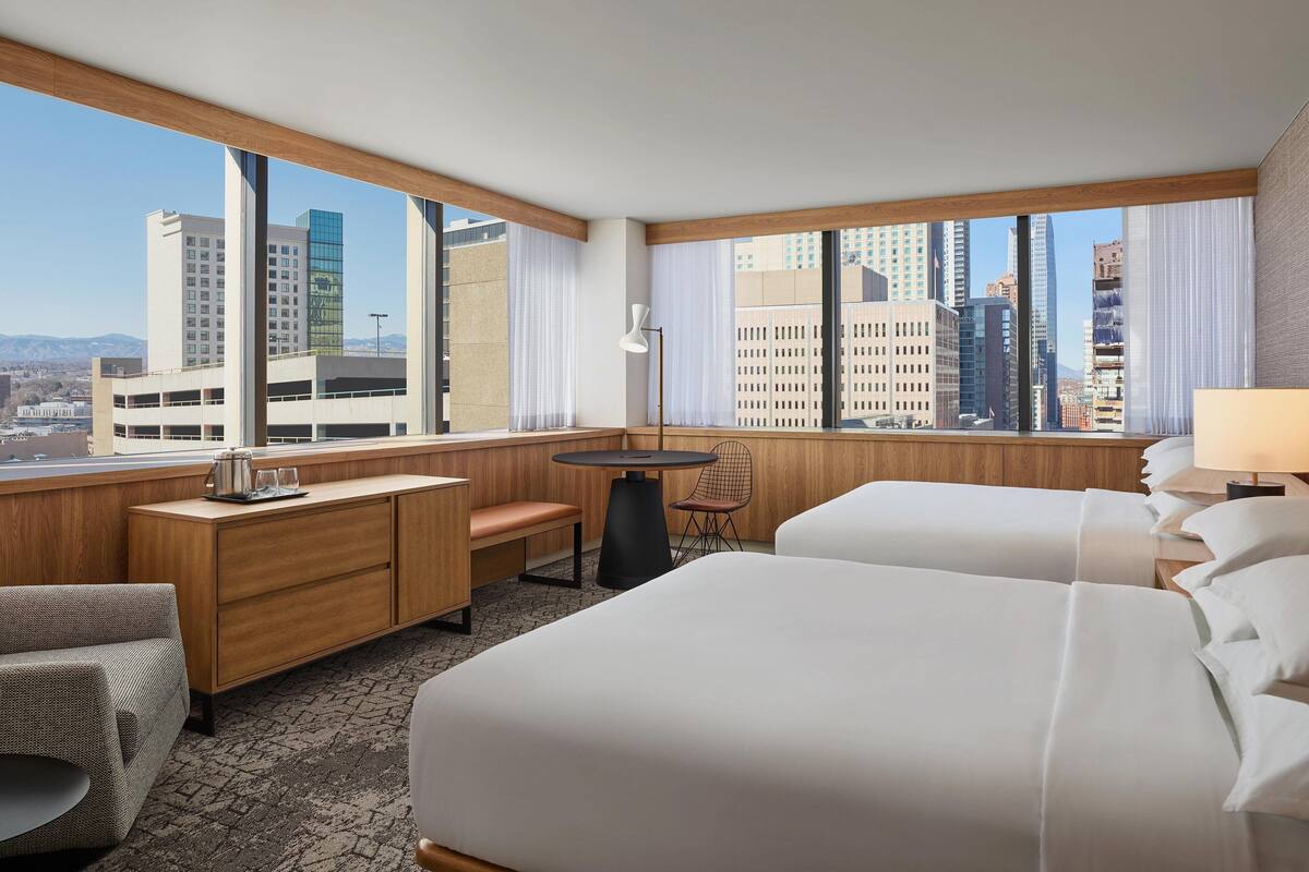 A hotel room with two beds, a desk, a chair, a small table, and large windows offering a city view.